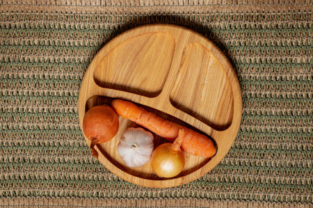 Wooden plate with onion, garlic and carrot on wicker tablecloth, natural vegetables for cooking at homeの写真素材