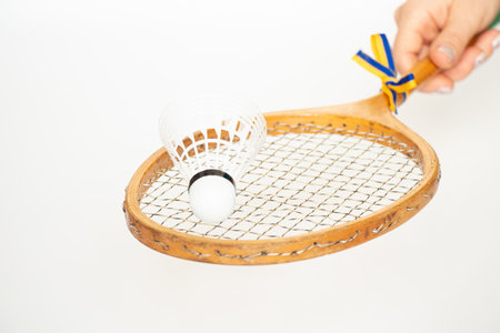 Female hand holding vintage racket with Ukrainian flag ribbon and shuttlecock on white background