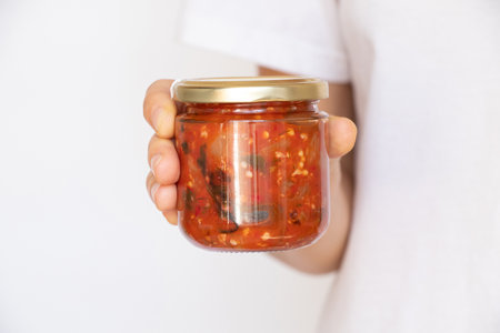 Homemade adjika in a glass jar with a golden lid in hand on a white background, canned hot sauce with herbs and spicesの写真素材