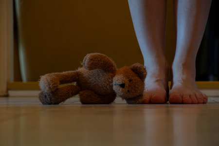 A child's teddy bear lies on the floor in the hallway of a house in the dark and next to it are women's legs, an abandoned teddy bearの写真素材