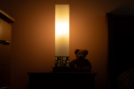 Teddy bear near a night lamp on a nightstand in a cozy bedroom with warm light near the bed, room lightingの写真素材