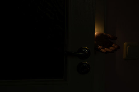 Female hand reaching through a half-open door with dim light in a dark room, concept of mystery, intrusion, help or fearの写真素材