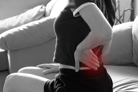 Girl holding her lower back in pain while sitting on the couch - acute back pain, back painの写真素材