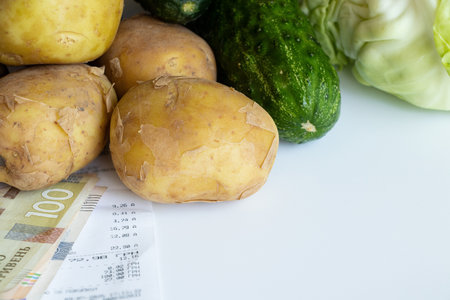 A check from a store and hryvnia lie on a white background next to vegetables, potatoes, cucumbers and cabbage, the price of vegetables in Ukraineの写真素材