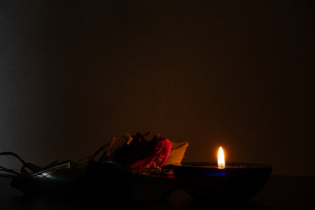 Burning candle and red rose lying next to it on table at home in the dark, candle flame