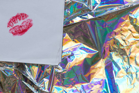 Kiss imprint with red lipstick on white paper lying on bright rainbow holographic film as a symbol...