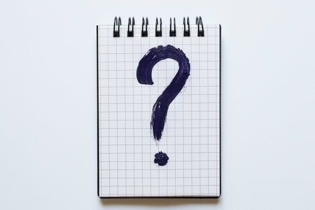 A black question mark drawn on a spiral notebook in a grid which lies on a white background