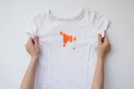 White T-shirt with orange paint or food stain on chest near pocket in hands of girl on washing theme, dirt, traces of food or childrens play on whiteの写真素材