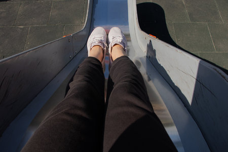 Legs of a child sitting on a metal slide, getting ready to slide down at a playground on a summer day in a parkの写真素材