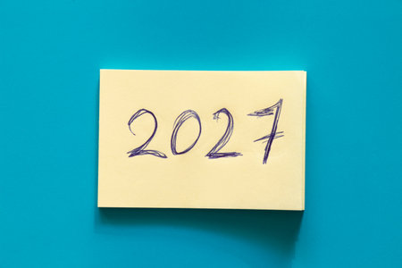 Yellow sticky notepaper with large handwritten 2027 inscription in blue ink on blue background on the theme of new year and plans, date and futureの写真素材