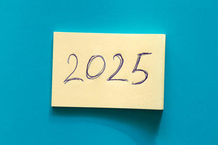 Yellow sticky notepaper with large handwritten 2025 inscription in blue ink on blue background on the theme of new year and plans, date and futureの写真素材