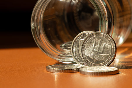 A 10 hryvnia coin with the coat of arms and the inscription State Special Transport Service placed on a brown leather surface and a glass jarの写真素材