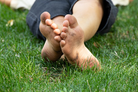 In summer on green grass in Ukraine little boy lies barefoot with dirty feet in parkの写真素材