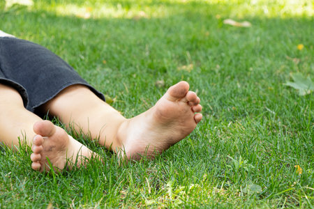 In summer on green grass in Ukraine little boy lies barefoot with dirty feet in parkの写真素材