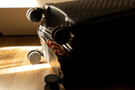 A black suitcase on wheels stands on the floor with a human leg sticking out of the half-open zipper.の写真素材