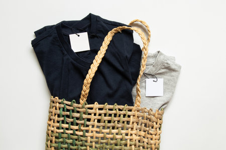 Two gray and blue t-shirts with tags lie in a straw bag on a white background as a mockup for design...