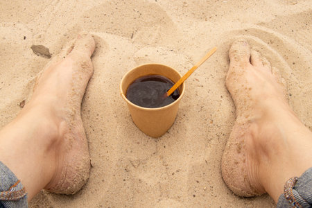 A paper cup with coffee and a wooden stick sits on the sand next to bare female feet in jeans,...