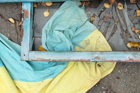 The Ukrainian flag lies on the asphalt among broken debris and an old wooden frame, symbolizing destruction, war, and the city's lost peaceの写真素材