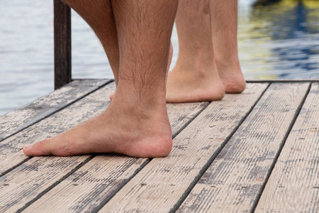 A mans bare, wet feet stand on old wooden pier planks covered in sand and moisture, creating the atmosphere of a summer vacation by the waterの写真素材