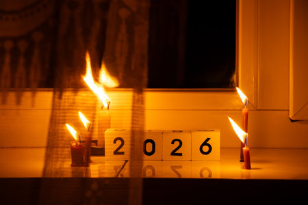 Cubes with the number 2026 and burning candles on a windowsill in a dark apartment, symbolizing life...