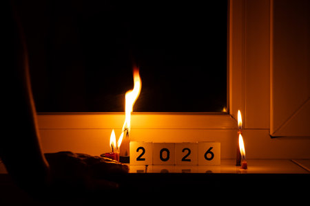 Cubes with the number 2026, burning candles, and women's hands on a windowsill in a dark apartment,...