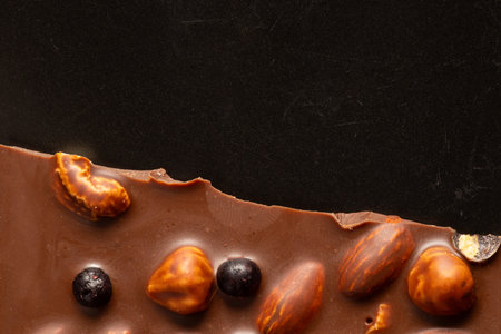 A bar of milk chocolate with almonds, hazelnuts, and whole berries on a black plate. The texture of...