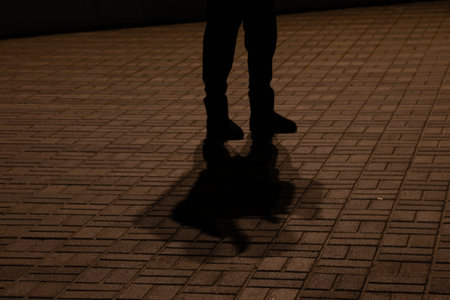 A man stands on a sidewalk in the dark at night. His feet are visible in shoes and a long shadow on...