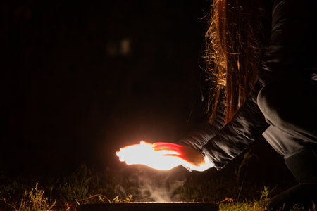 A girl warms her hands near a bright lamp on the ground in a dark winter park. A cold night. An...
