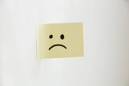 A yellow sticky note with a sad face drawn on it hangs on a white background, depicting a bad mood, sadness, and negative emotionsの写真素材