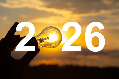 A hand holding a light bulb against a golden sunset with a large number 2026, a symbol of energy,...