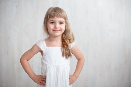 Portrait of pretty little girl in white dress , looking at camera and smiling, standing against gray background. She is happy and waiting for a moment.の写真素材