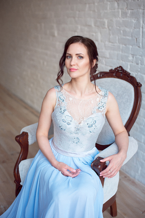 Beautiful luxurious woman with medium brown hair in a long light blue dress sits on an armchair in the room.の写真素材