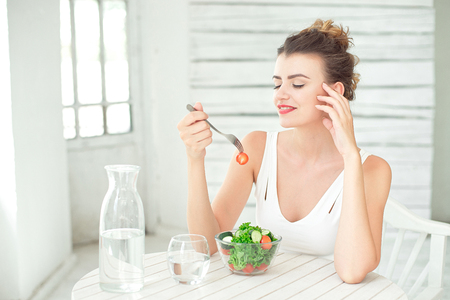 Portrait of a young smiling woman eating a fresh salad in white room.の写真素材