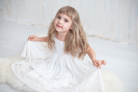 Portrait of pretty little girl in white wide dress sitting on the floor. Blonde girl 4-5 years indoor.の写真素材