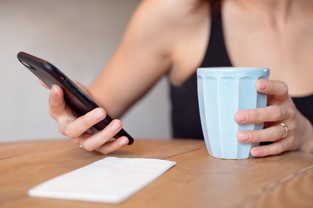 Female hands holding cell phone and a cup of coofee on the wooden table.の写真素材