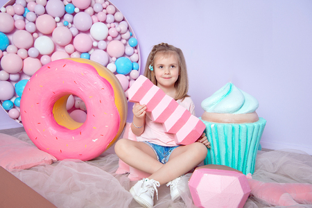 Little girl sitting in colorful room around big toyful candies, donut, lollipopの写真素材