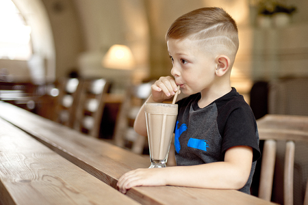 little boy drinking milkshake sitting at the table in cafeの写真素材