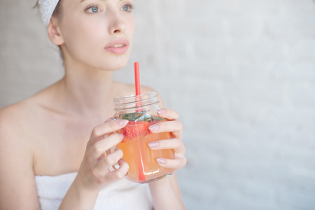 Caucasian smiling woman after spa covered with white towel drinking refreshing drink in glass bottle through strawの写真素材