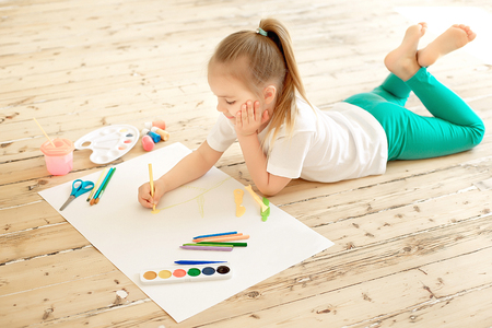 Top view of little blonde girl painting on big white paper while laying on the floor indoorsの写真素材