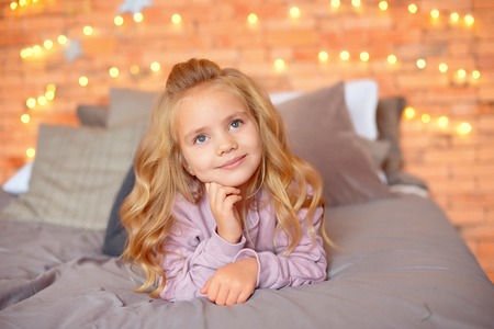 Beautiful girl with blond curls in expectation of gifts in the background Christmas lights and bed . New Year and Christmas conceptの写真素材