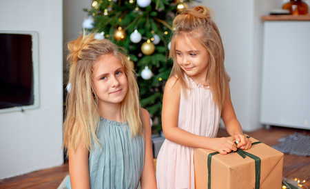 Two sisters with blond hair in luxurious dresses are sitting near the Christmas tree and looking at the gifts. New Year and Christmas Conceptの写真素材