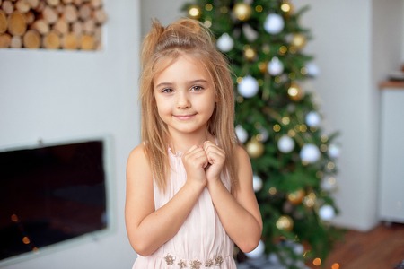 Happy little girl in luxury dress holds hands near face.With colorful lights and Christmas tree on background. Holidays, christmas, new year, x-mas concep.の写真素材