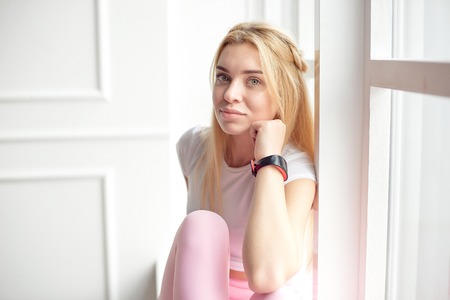 Young slim woman with an athletic body with long blond hair, dressed in white sportswear and pink leggings, sits on the floor in a bright yoga room with a large panaramic window, workout program for the gymの写真素材
