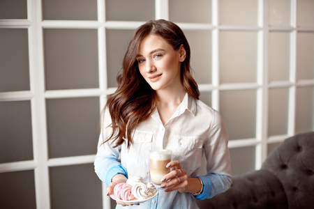 Beautiful young woman with dark hair dressed in a white shirt is standing in a cafe indoors with a glass cup of latte on soy milkの写真素材