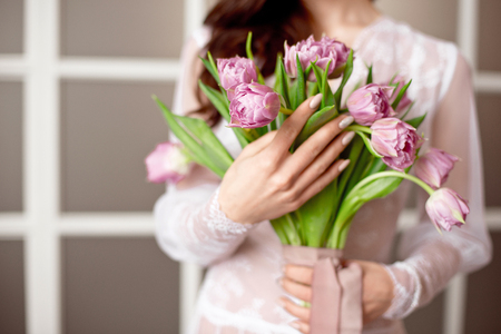 Beautiful young woman in the white dress with flowers tulips in hands on bed at homeの写真素材