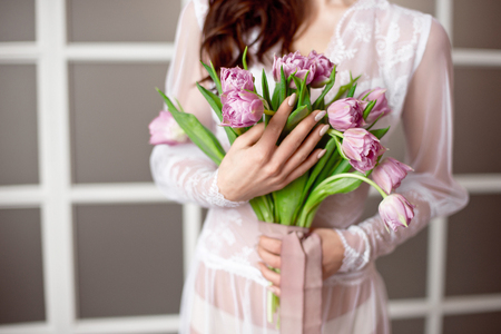 Beautiful young woman in the white dress with flowers tulips in hands on bed at homeの写真素材