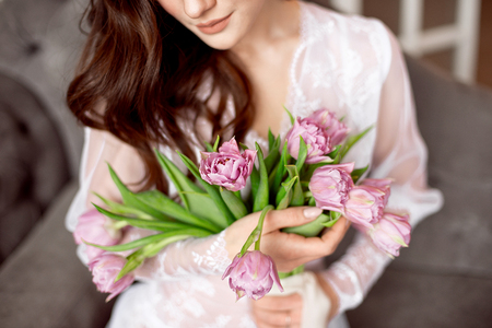 Beautiful young woman in the white dress with flowers tulips in hands on bed at homeの写真素材