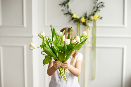 Portrait of a pretty little girl in a white shirt hiding behind a bouquet of white tulips.の写真素材