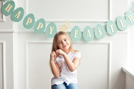 Cute little blond girl sitting in a bright room near the window on the background of streamers with the inscription Happy Easter.の写真素材