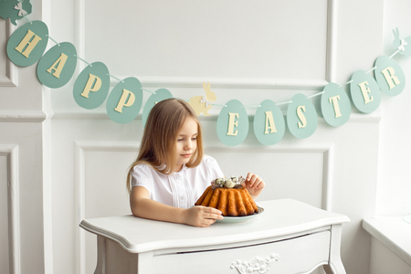 Cute little blonde girl is sitting at a white vintage table and looking at a tasty cake with interest.の写真素材